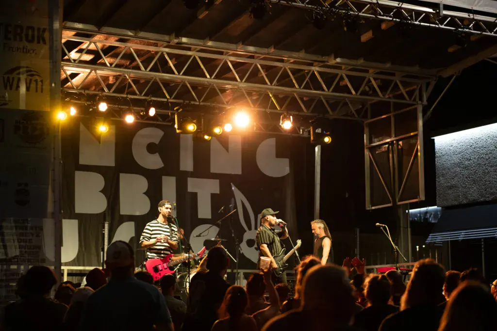 A four-piece band performs on an illuminated stage before a crowd at an outdoor music festival.