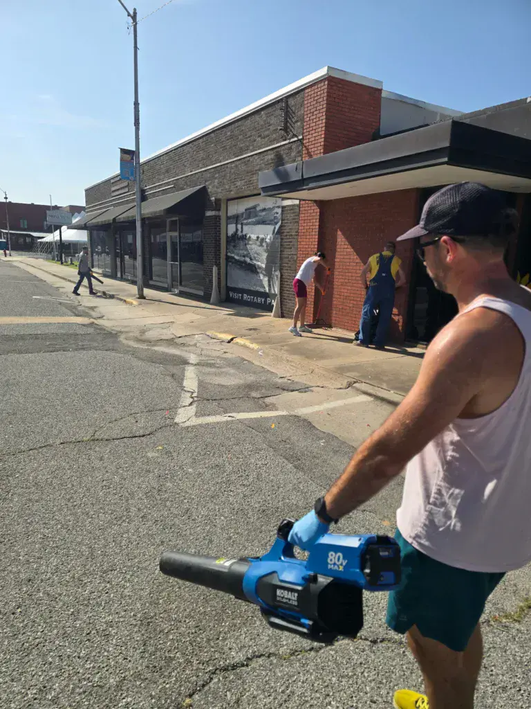 A person holding a blue power tool stands on a street outside brick buildings during a festival.