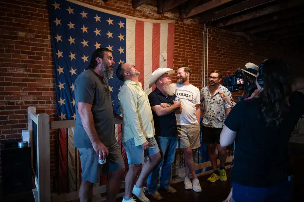 A group of people gather indoors in front of an American flag while someone records video.