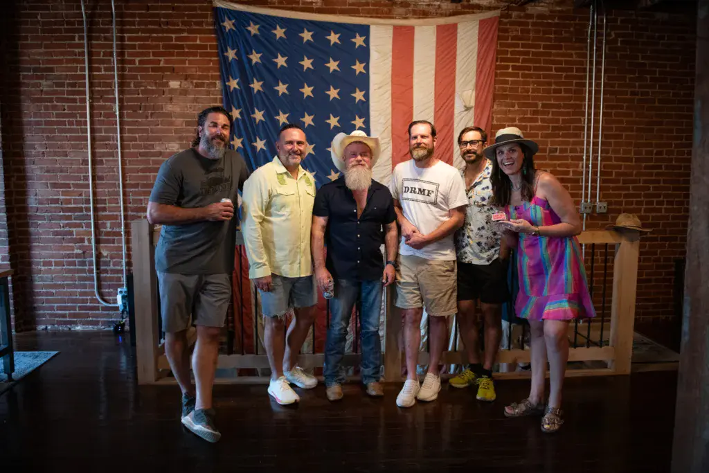 Six people stand together on a stage in front of an American flag backdrop inside a brick venue.