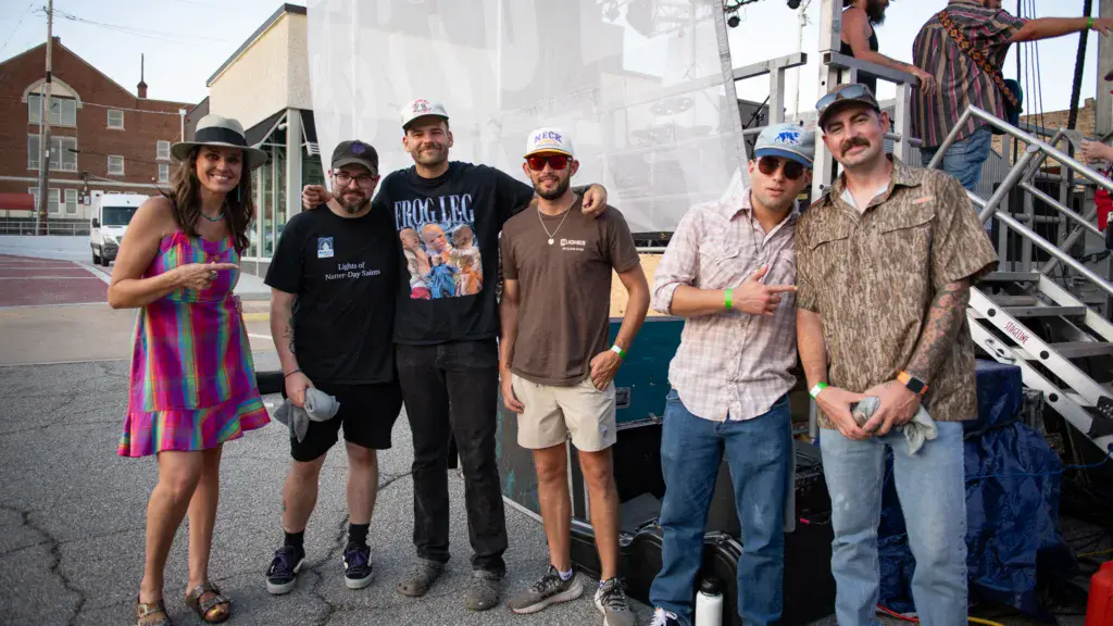 A group of six people stand together in front of a stage setup at an outdoor music festival venue.