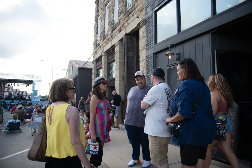 Festival attendees gather in a group outside brick buildings at an outdoor music venue.