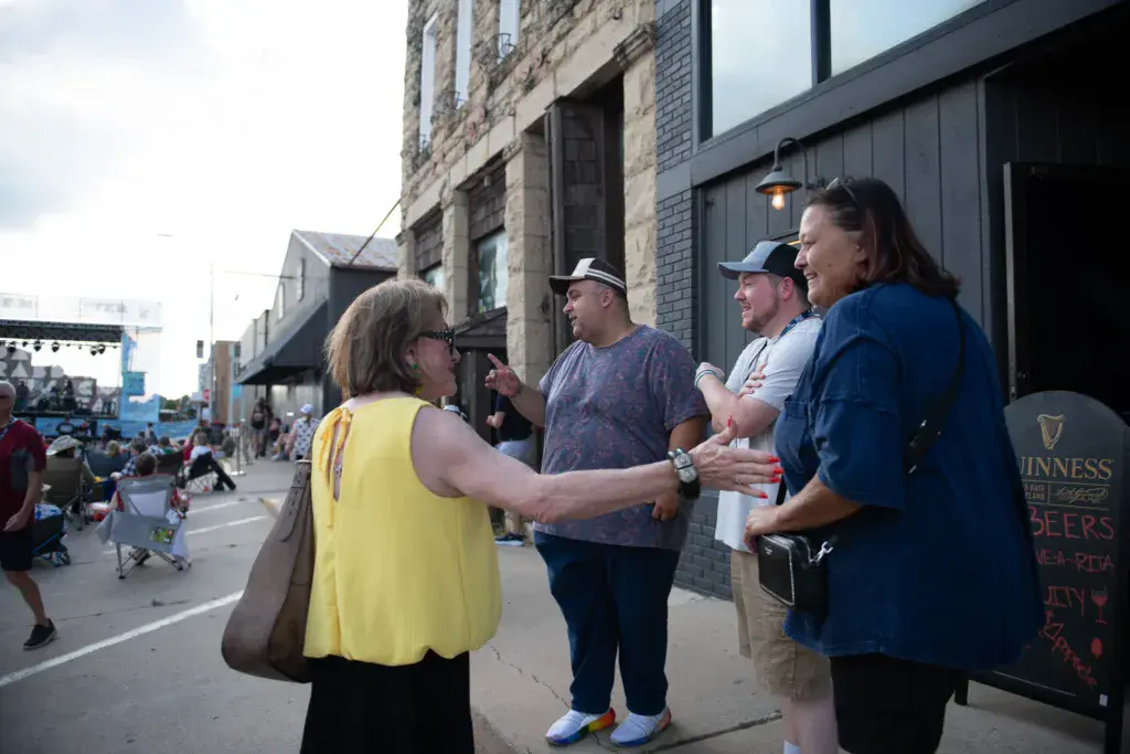 A festival attendant in a yellow vest speaks with a group of visitors outside a brick building at an outdoor music festival.
