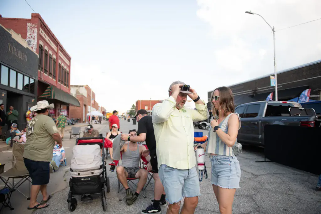 Festival attendees enjoy a sunny day on a street lined with historic brick buildings and parked vehicles.