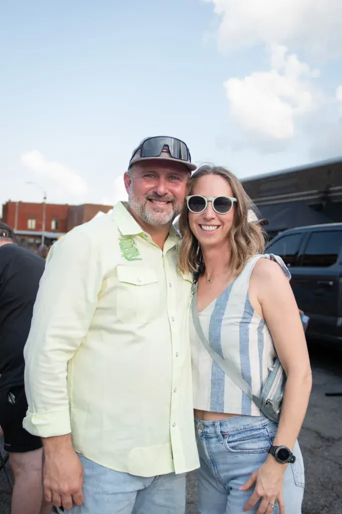 Two attendees smile together at the Dancing Rabbit Music Festival with vehicles and buildings visible in the background.