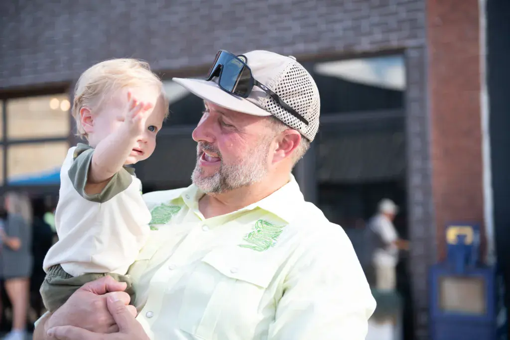 An adult wearing a white cap holds a young child at an outdoor festival near a brick building.