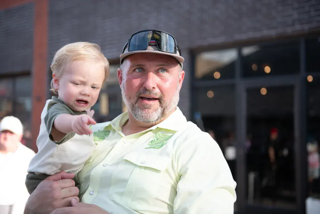 A man wearing a light green shirt and cap holds a toddler while standing outdoors at a festival venue.