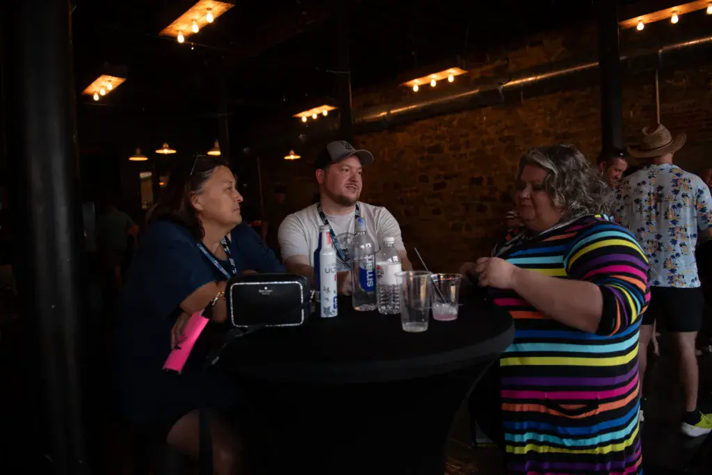 Festival attendees wearing lanyards gather around a table at an evening event under warm string lighting.