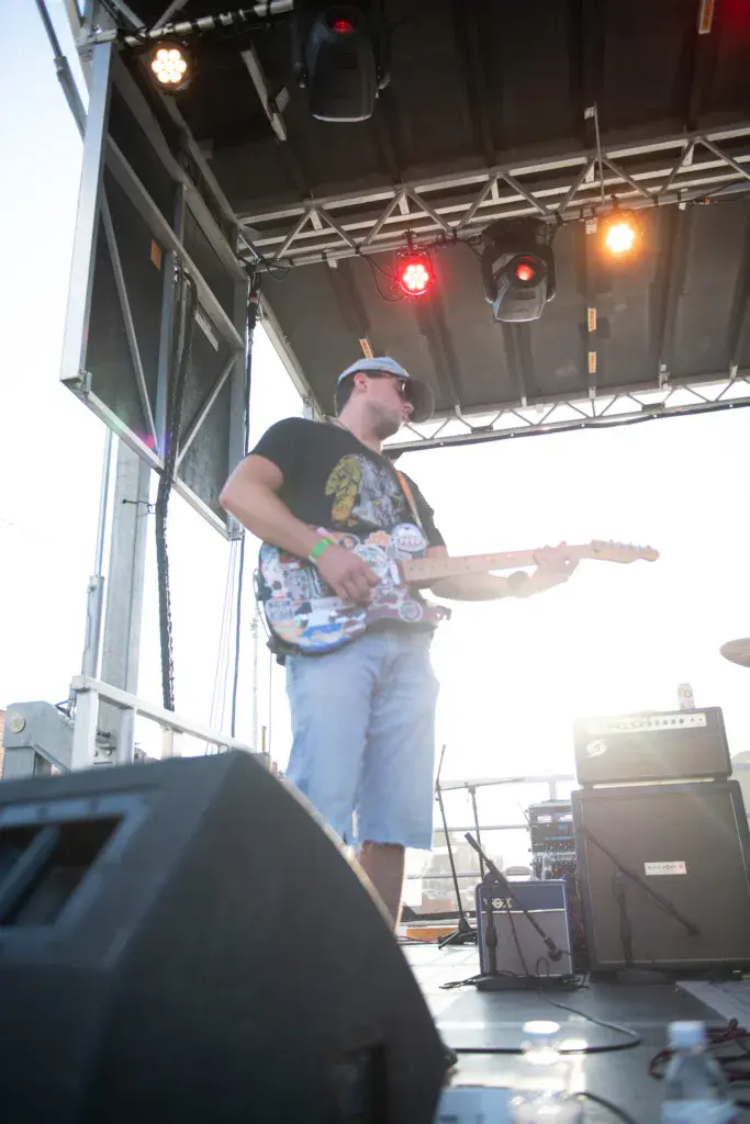Guitarist performing on an outdoor festival stage under stage lighting and metal rigging.