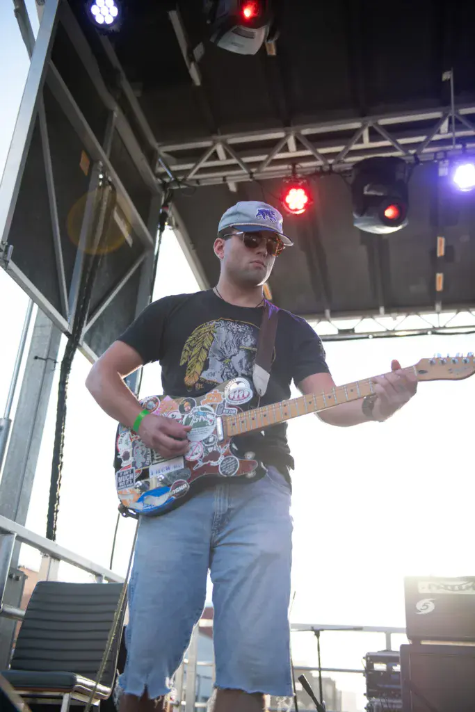 Guitarist performing on stage at night wearing a blue cap and decorated electric guitar under colorful stage lighting.