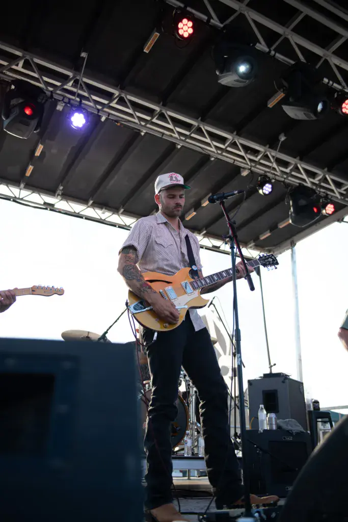 A guitarist performs on stage under colorful stage lights at an outdoor music festival.