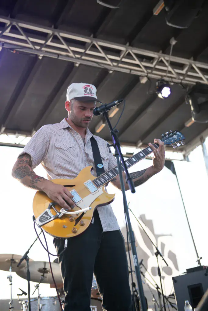 A guitarist performs at a covered stage, singing into a microphone while playing a yellow electric guitar.
