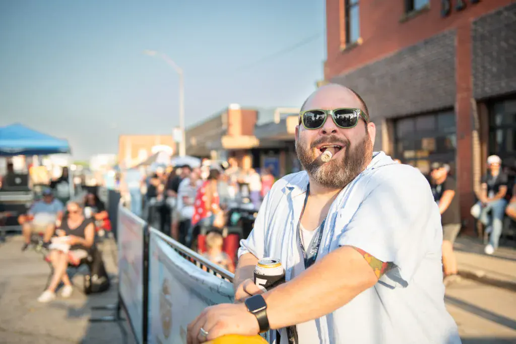 A bearded man wearing sunglasses and a light blue shirt smiles while holding a drink at an outdoor festival venue.
