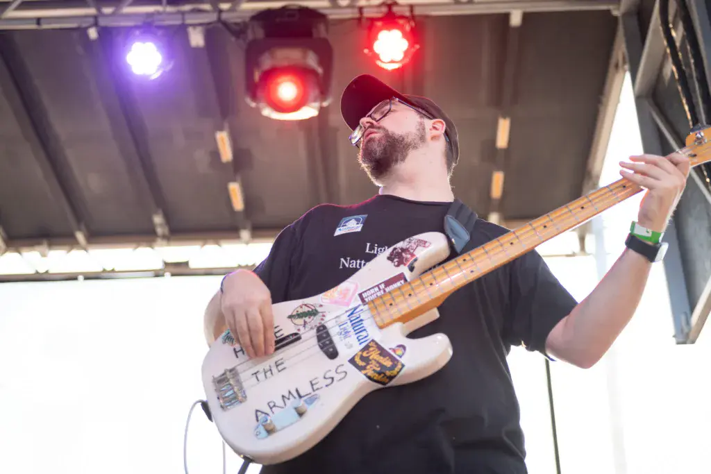 A bass guitarist wearing glasses and a maroon cap performs on stage under colorful stage lighting.