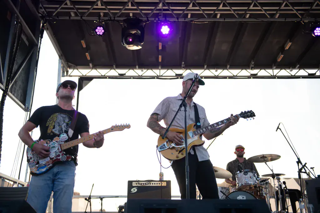 Three musicians perform on stage under bright purple stage lights during a daytime outdoor music festival.