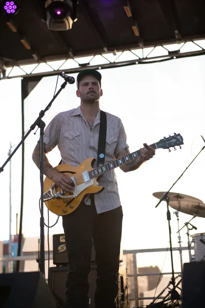 A guitarist performs on stage under a tent canopy at an outdoor music festival.
