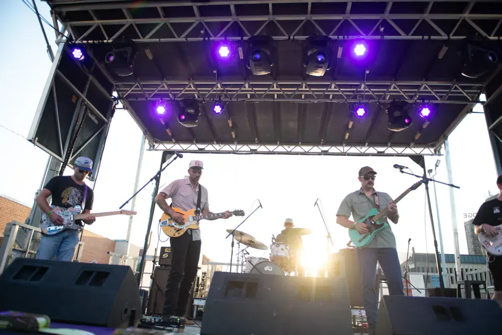 A band of four musicians performs on an outdoor stage with purple lighting at sunset.