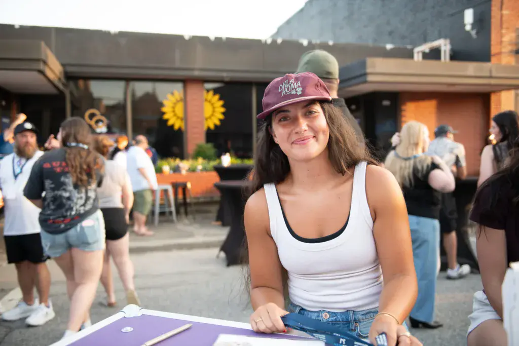 A young woman in a maroon cap smiles at the camera while holding a purple item at an outdoor festival venue.