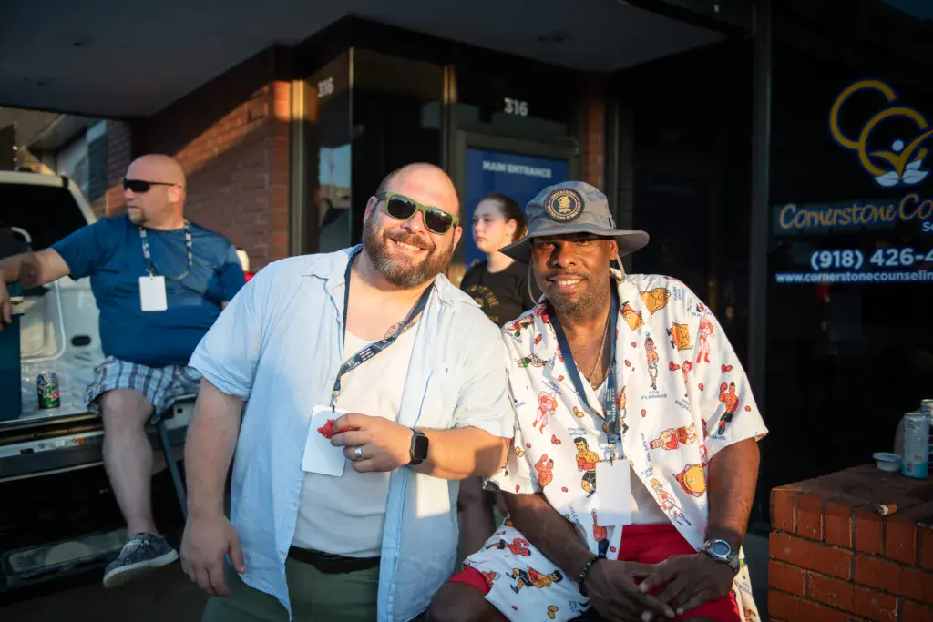 Two men wearing lanyards smile at the Dancing Rabbit Music Festival venue, with others visible in the background.