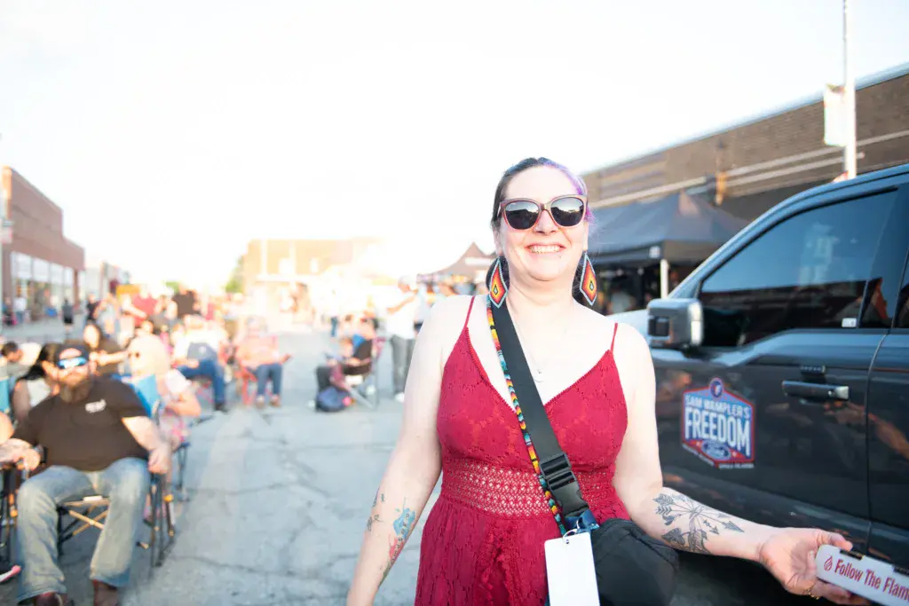 A smiling woman in a red dress and sunglasses stands in front of festival crowds and parked vehicles.