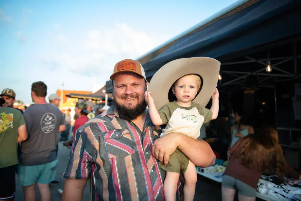 A smiling adult wearing a striped shirt and cap holds a young child in a large white cowboy hat at an outdoor festival.