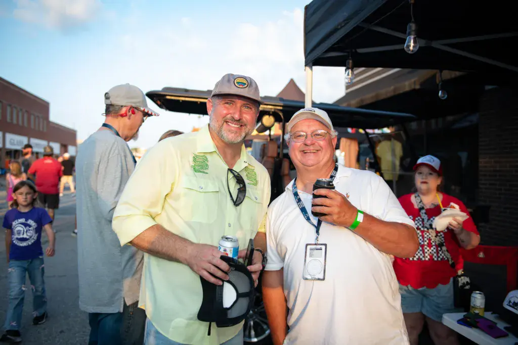 Two men smile together at an outdoor festival venue under a black canopy structure.