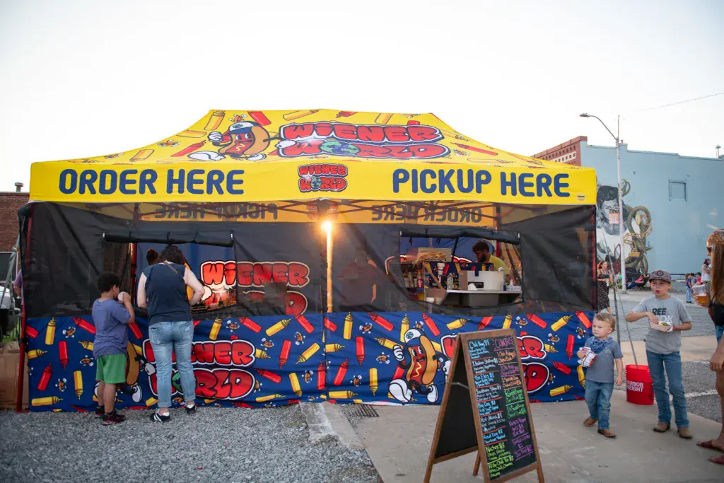 A bright yellow food vendor tent with hot dog and wiener branding serves customers at an outdoor festival.