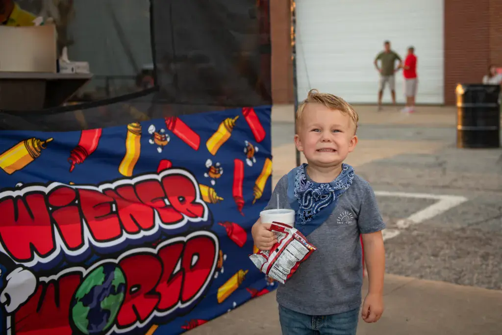 A young child wearing a blue bandana and gray shirt smiles at the camera while holding a drink at an outdoor music festival venue.