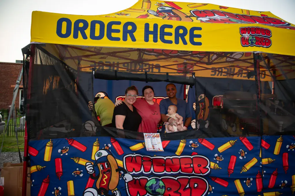 Staff members smile at a colorful food vendor booth with yellow canopy at the festival.