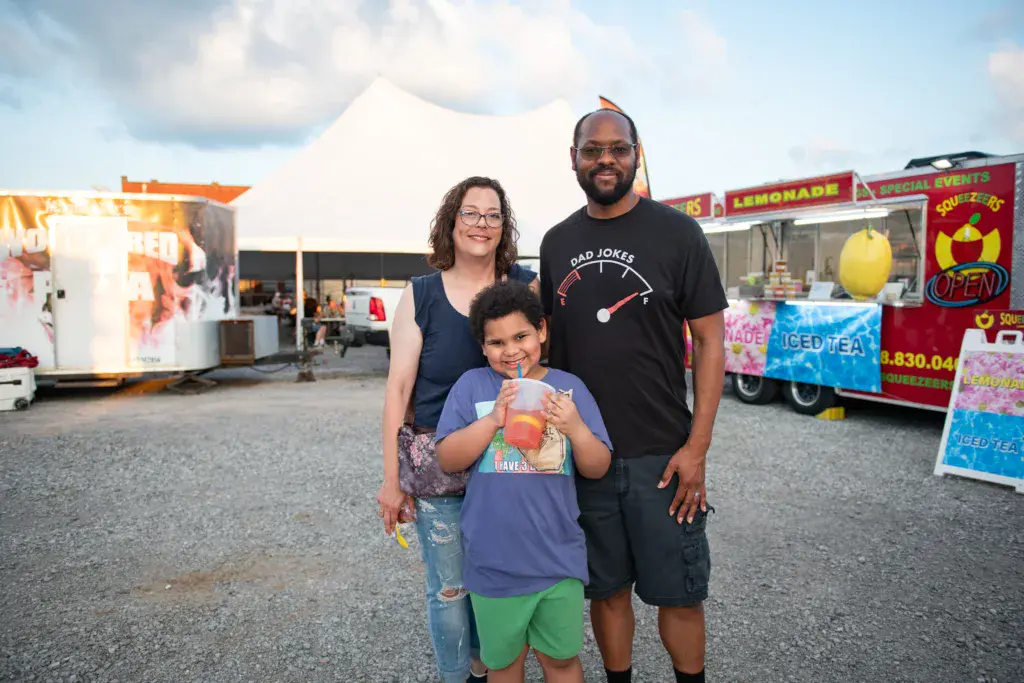 Family of three smiling at a festival food vendor area with food trucks and concession stands in the background.