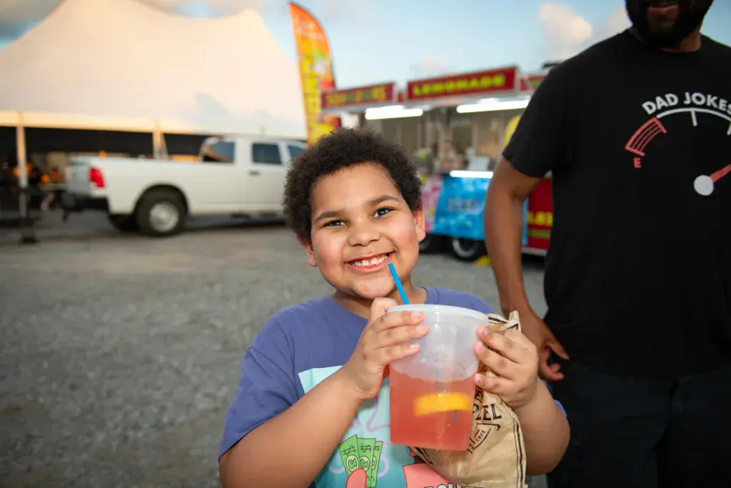 A smiling child holds a beverage at a festival with vendor booths and vehicles visible in the background.