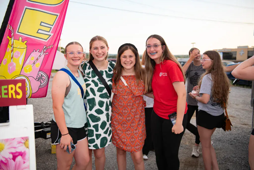 A group of smiling young people pose together at an outdoor festival venue with a colorful pink food truck visible in the background.