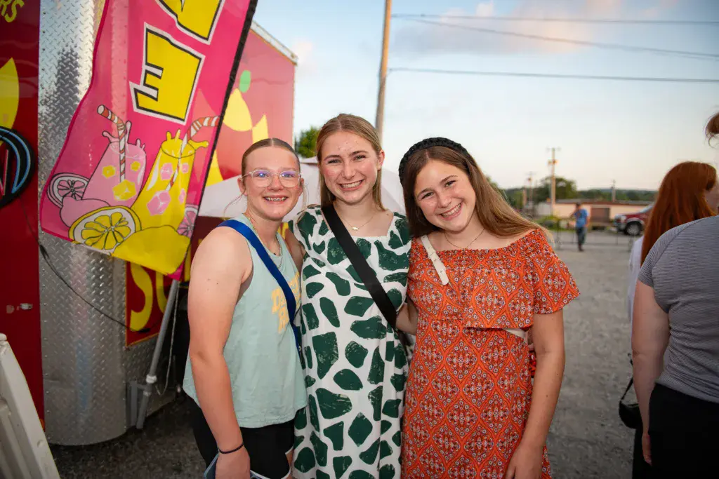 Three smiling young women posing together at an outdoor festival venue with colorful signage behind them.