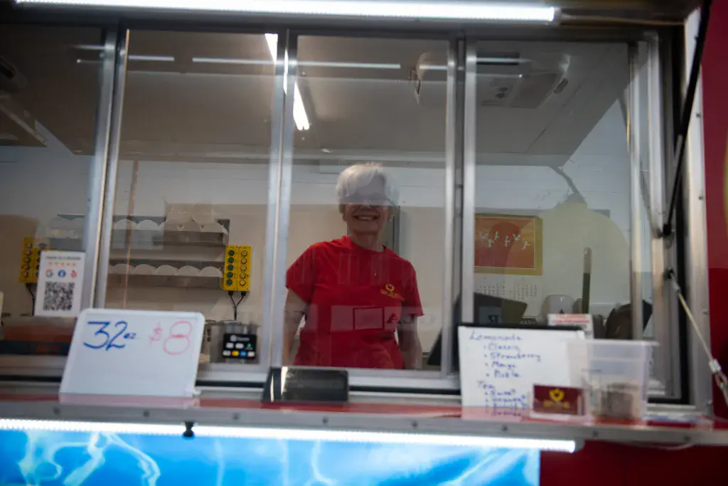 A vendor in a red shirt stands inside a food service window with price signage and menu items visible.