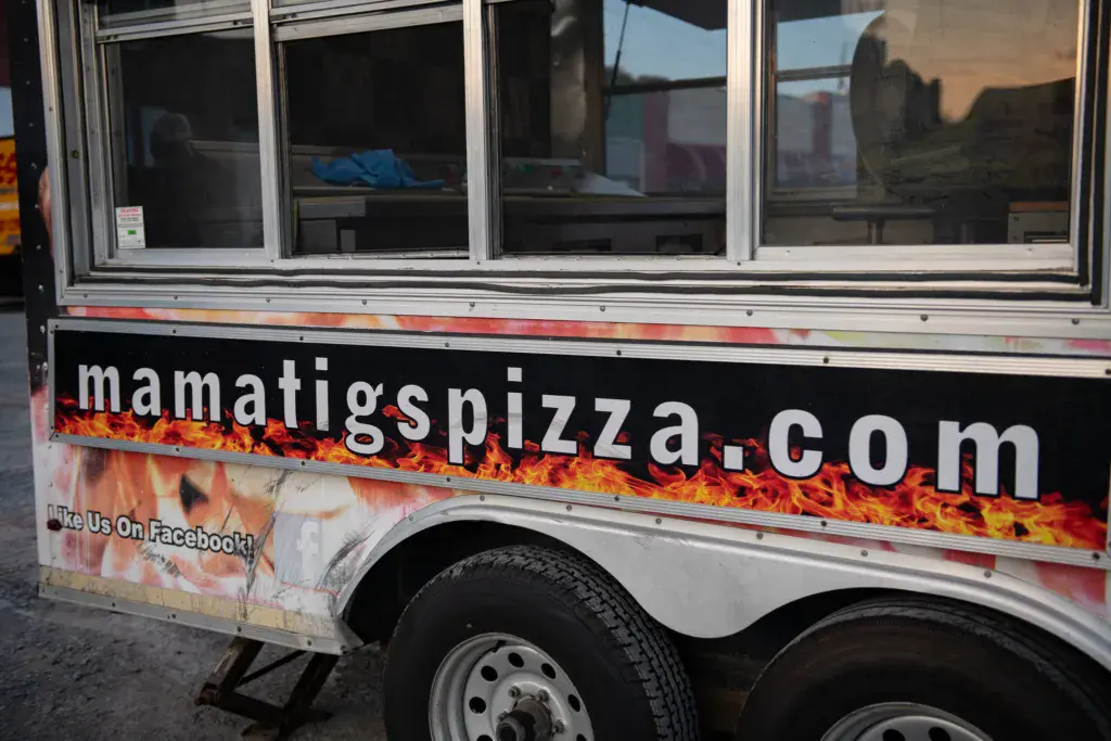 A food truck with flame graphics displays pizza website and Facebook information.