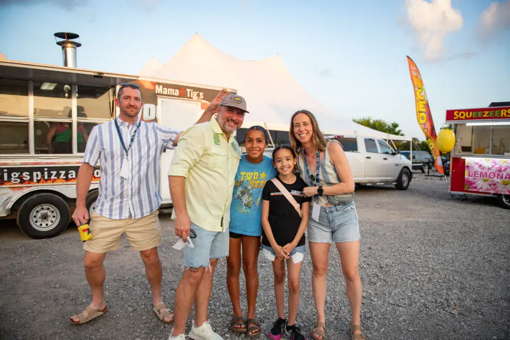 A group of five people pose together at a festival with food vendor trailers and a white tent visible in the background.