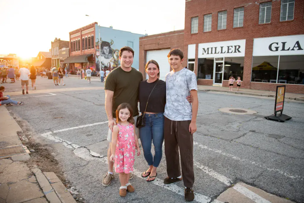 A family of four smiles at the camera while standing on a street with historic downtown buildings and festival crowds in the background.