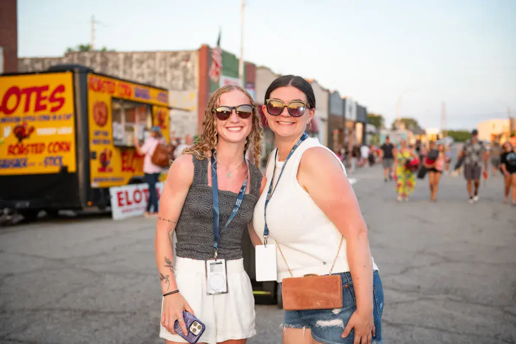 Two people wearing sunglasses and festival lanyards smile at the camera in front of food vendor booths and crowds at an outdoor festival.
