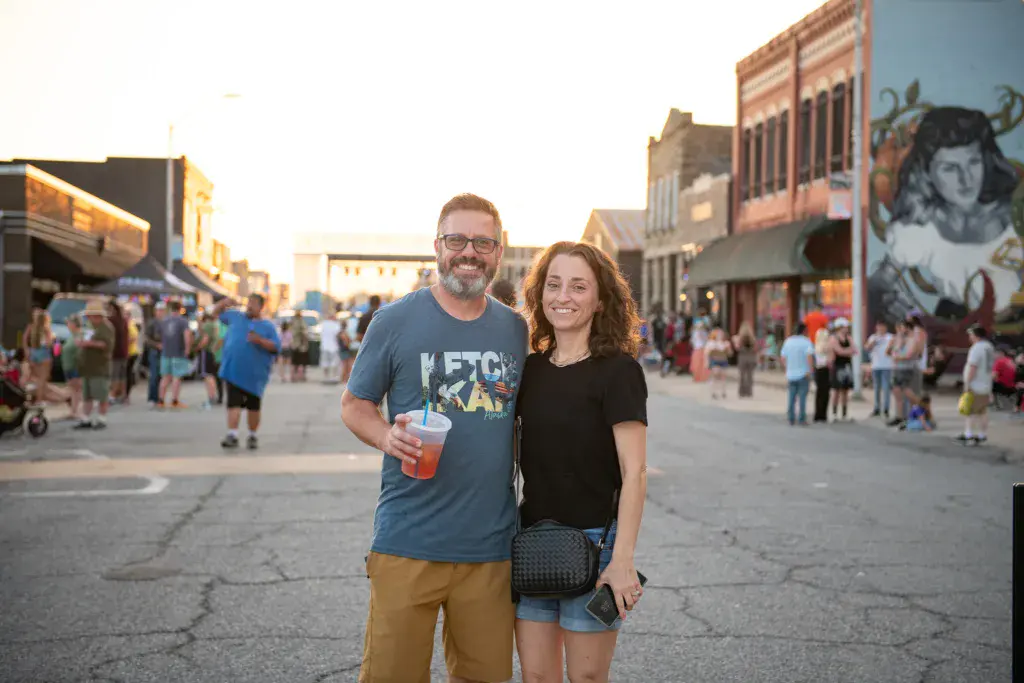 Two people smile at the camera in a festival street lined with historic brick buildings and crowds.