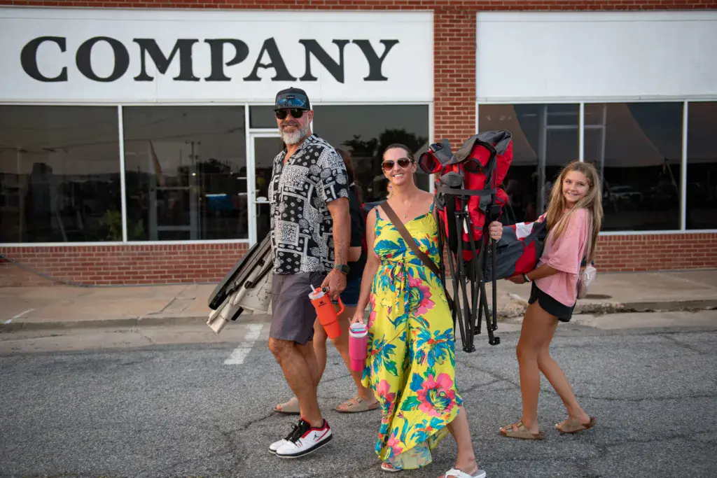 Three people carrying bikes and equipment stand in front of a brick building with a company sign.