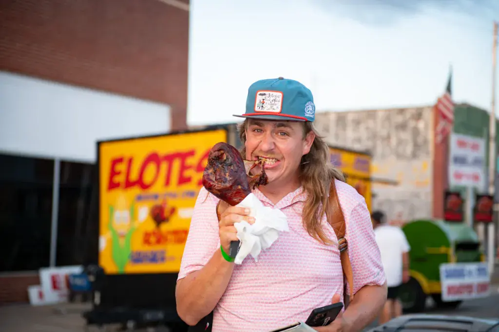 A person in a pink shirt and blue cap holding a large corn dog at an outdoor festival with food vendor signage in the background.