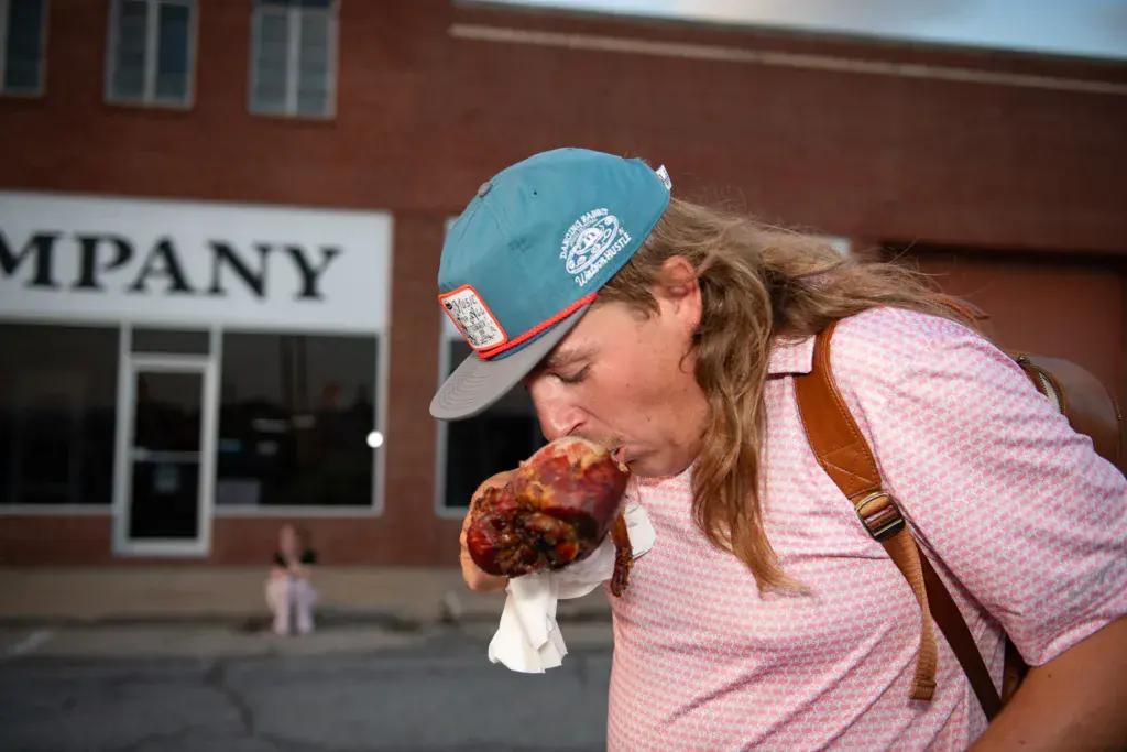 A person in a blue cap and pink shirt eats food while standing in front of a brick building with a storefront.