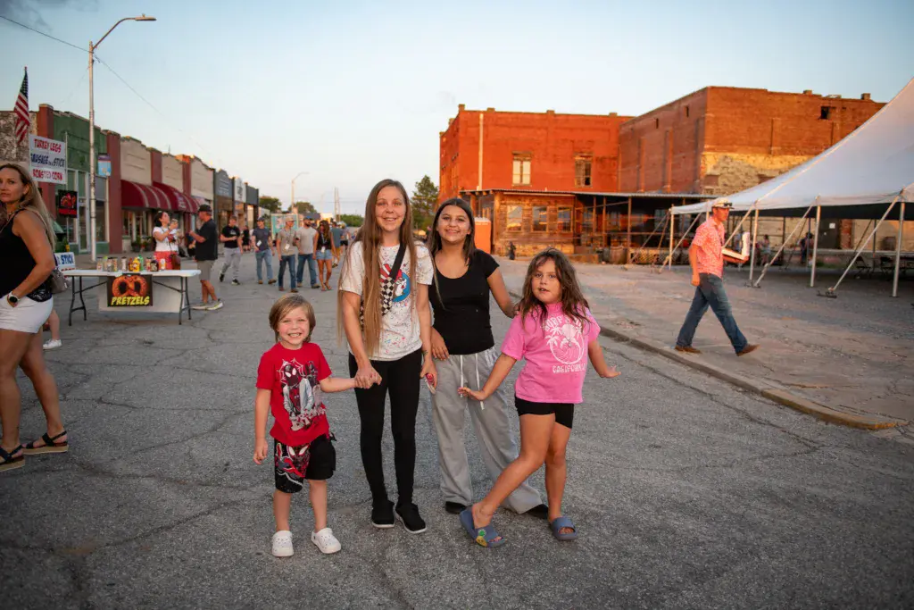 An adult with three children stands together in a festival venue in front of historic brick buildings and vendor tents.
