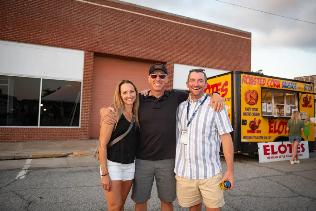 Three people pose together in front of a brick building and a food vendor stand at the festival.