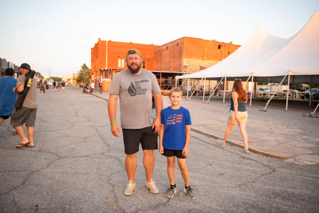 An adult and child stand together in the festival grounds with an orange building and white tent structures in the background.