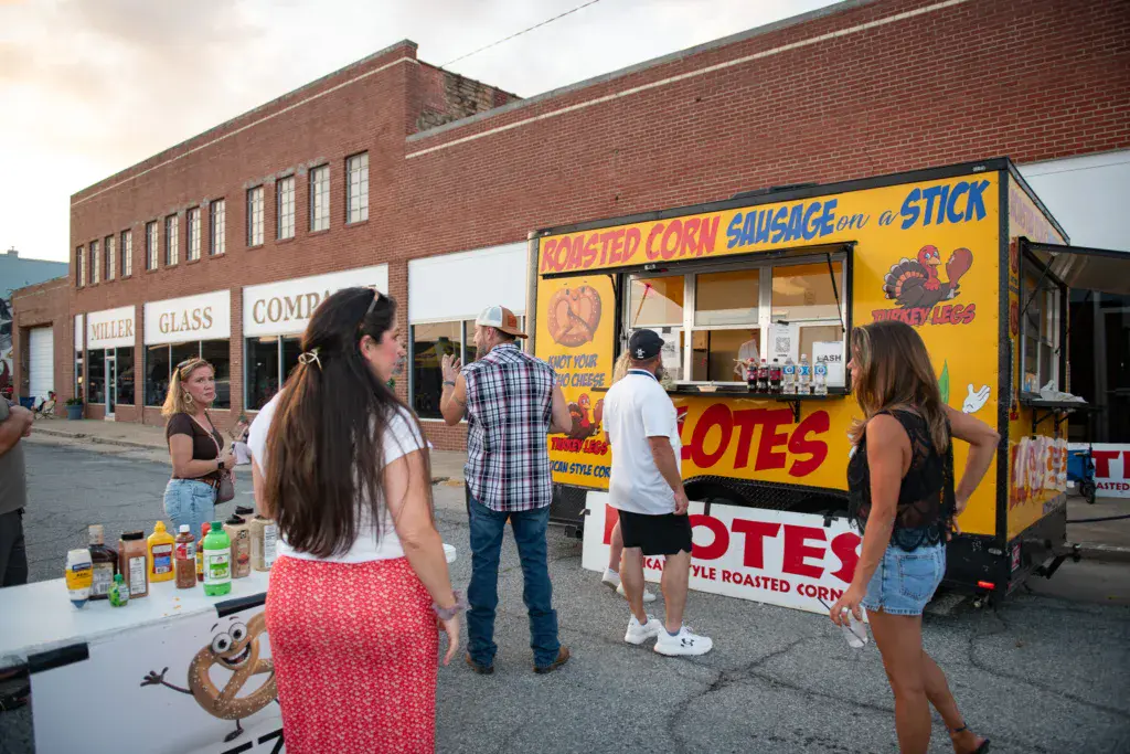 Festival attendees gather around a yellow food vendor truck selling roasted corn and sausage near a brick building.