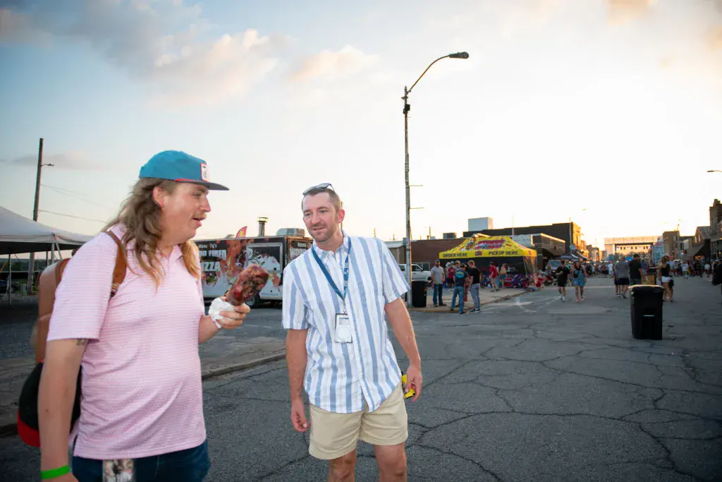 Two people walk and chat in a festival venue with crowds and a stage visible in the background.