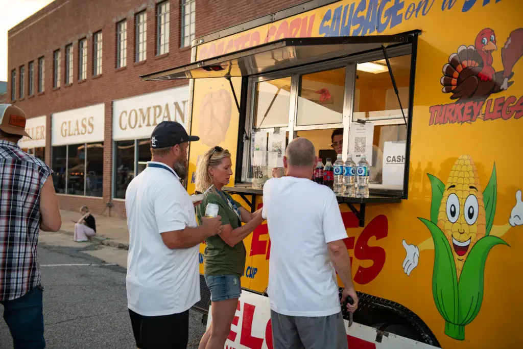 Festival attendees wait in line at a yellow food truck with colorful painted graphics.