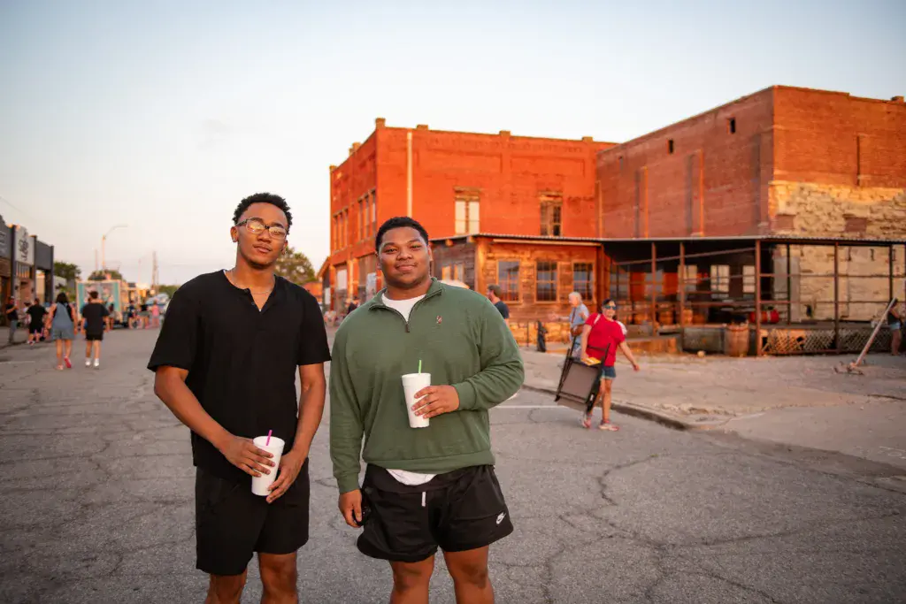 Two attendees stand holding drinks in front of a large orange brick building at the festival venue.