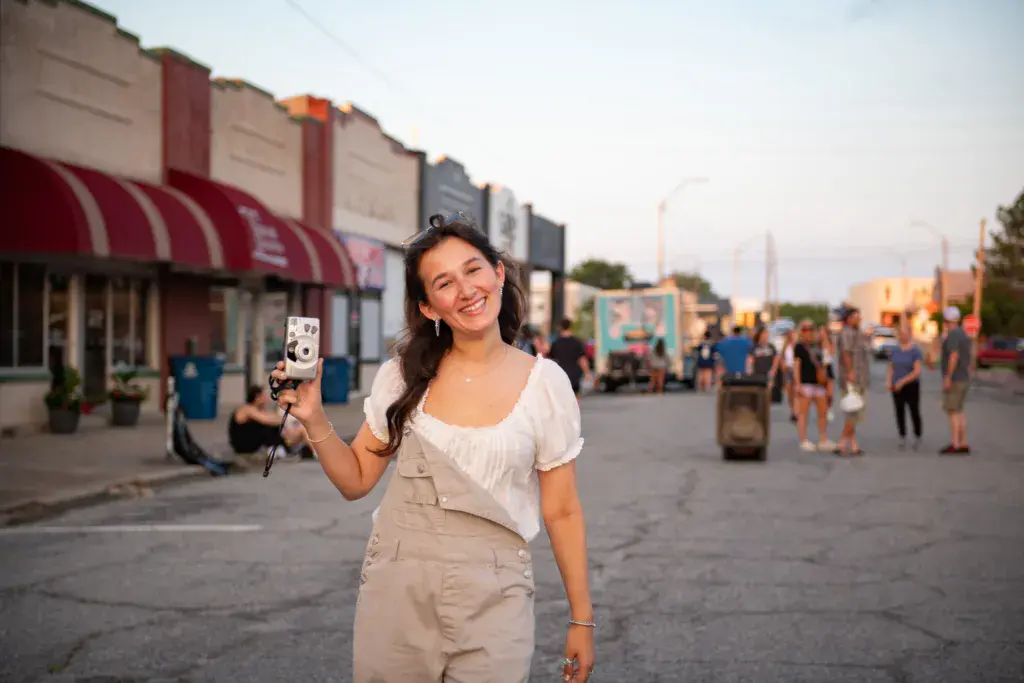 A person in a white blouse and tan vest holds a vintage camera while smiling at an outdoor festival venue.